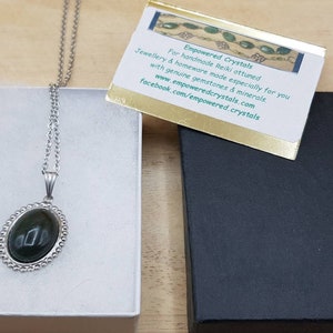 May include: A silver chain necklace with a dark green oval pendant set in a silver bezel. The pendant is in a white box with a black box next to it. The black box has a gold card on top with the text "Empowered Crystals" and information about the company.