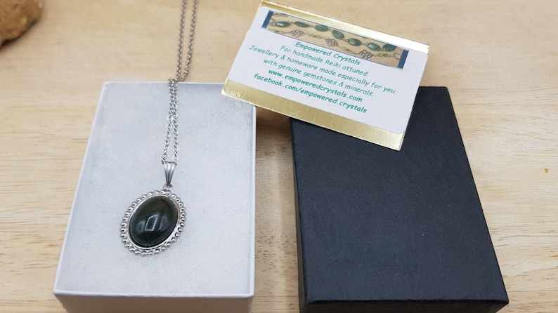 May include: A silver chain necklace with a dark green oval pendant set in a silver bezel. The pendant is in a white box with a black box next to it. The black box has a gold card on top with the text "Empowered Crystals" and information about the company.