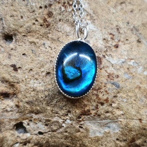 May include: A silver necklace with an oval pendant. The pendant features a vibrant blue and green iridescent stone, set in a silver bezel. The necklace is displayed on a textured, light brown stone surface.