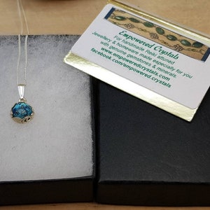 May include: A silver necklace with a blue and green glass pendant in a black box. The box has a card on top that reads "Empowered Crystals" and includes the website and Facebook page for the company.