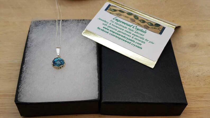 May include: A silver necklace with a blue and green glass pendant in a black box. The box has a card on top that reads "Empowered Crystals" and includes the website and Facebook page for the company.