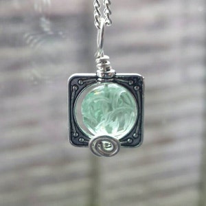 May include: A silver pendant necklace with a square frame and a green glass orb inside. The orb has a swirling pattern.