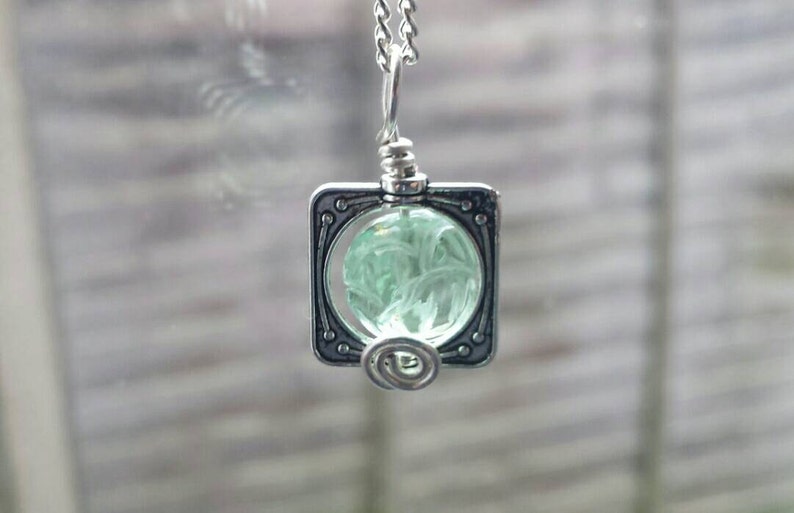 May include: A silver pendant necklace with a square frame and a green glass orb inside. The orb has a swirling pattern.