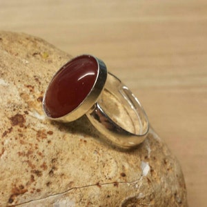 May include: A silver ring with a red carnelian gemstone. The ring is set on a light brown rock.