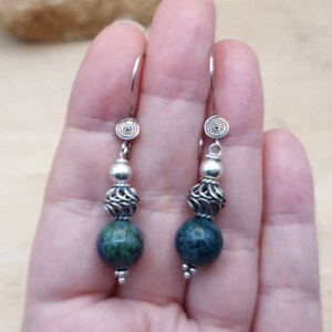 May include: A pair of dangle earrings with a spiral design at the top. The earrings feature a silver bead, a filigree bead, and a dark blue and green stone. The earrings are suspended from a silver hook.