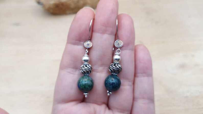 May include: A pair of dangle earrings with a spiral design at the top. The earrings feature a silver bead, a filigree bead, and a dark blue and green stone. The earrings are suspended from a silver hook.