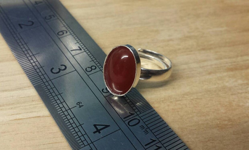 May include: A silver ring with a red oval-shaped gemstone. The ring is shown next to a ruler, with the gemstone measuring approximately 1 inch in length.