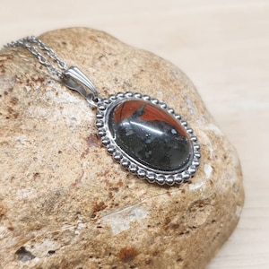 May include: A silver chain necklace with a black and red oval pendant. The pendant has a beaded border and a black stone with red accents.