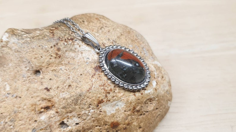 May include: A silver chain necklace with a black and red oval pendant. The pendant has a beaded border and a black stone with red accents.