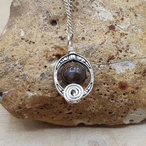 May include: A silver pendant necklace with a dark brown stone set in a circular wire frame. The pendant is on a silver chain and is resting on a brown rock.
