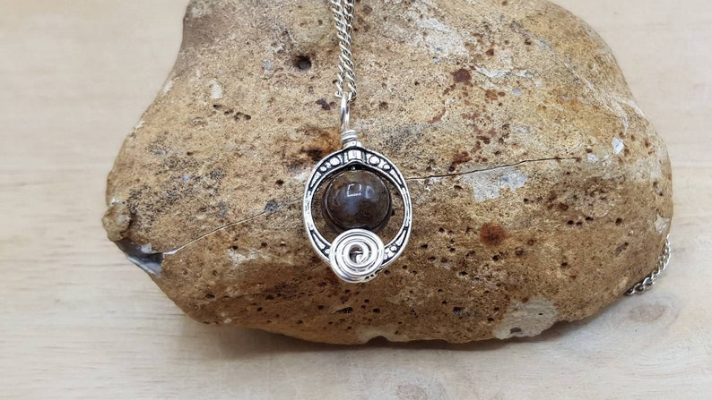 May include: A silver pendant necklace with a dark brown stone set in a circular wire frame. The pendant is on a silver chain and is resting on a brown rock.