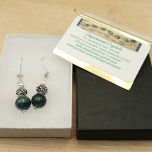 May include: A pair of handcrafted earrings with dark blue and silver beads, presented in a white box. The earrings feature a combination of round, textured beads and silver accents. A business card with text is also visible.