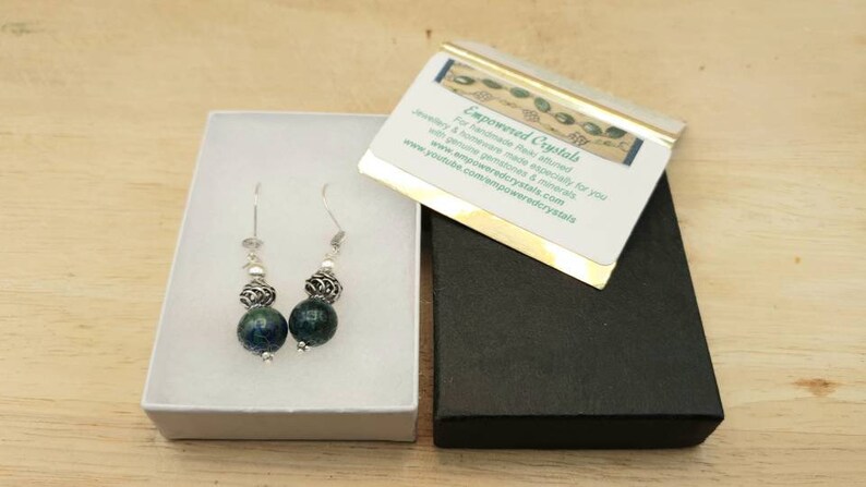 May include: A pair of handcrafted earrings with dark blue and silver beads, presented in a white box. The earrings feature a combination of round, textured beads and silver accents. A business card with text is also visible.