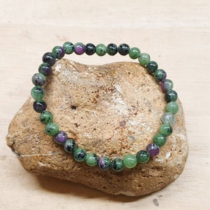 May include: A bracelet made of round beads in shades of green, purple, and black. The bracelet is displayed on a textured, light brown stone, with a light wood grain backdrop. The beads are evenly sized and spherical.