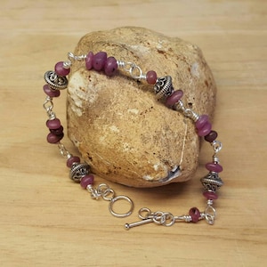 May include: A silver bracelet with a toggle clasp features pink and purple gemstone beads and silver accents.