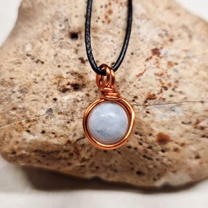 May include: A round, pale blue gemstone pendant secured in a copper wire wrap setting. The pendant hangs from a thin black cord. The wire is coiled tightly around the base of the stone and forms a small loop at the top for the cord to pass through.