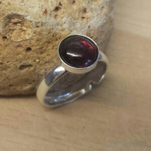 May include: A silver ring with a large, round, dark red gemstone. The ring has a simple, elegant design with a polished finish. The gemstone is set in a bezel setting. The ring is displayed on a light brown wooden surface.