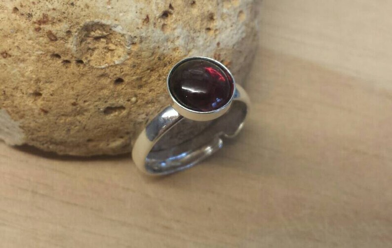 May include: A silver ring with a large, round, dark red gemstone. The ring has a simple, elegant design with a polished finish. The gemstone is set in a bezel setting. The ring is displayed on a light brown wooden surface.