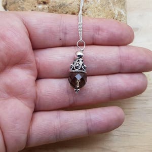 Op de afbeelding: Een zilveren ketting met een hanger met een gefacetteerde, rookkwarts edelsteen. De hanger heeft een decoratieve zilveren dop en een kleine zilveren kraal aan de onderkant. De ketting wordt op een hand getoond.