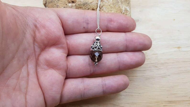 Op de afbeelding: Een zilveren ketting met een hanger met een gefacetteerde, rookkwarts edelsteen. De hanger heeft een decoratieve zilveren dop en een kleine zilveren kraal aan de onderkant. De ketting wordt op een hand getoond.