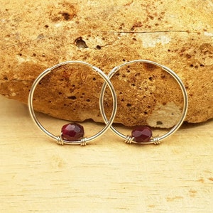 May include: A pair of silver hoop earrings with a single red gemstone bead on each hoop.