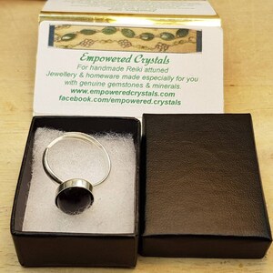 May include: A silver ring with a black gemstone, presented in a black box with a white interior. The box is labelled "Empowered Crystals" with a website and Facebook page.