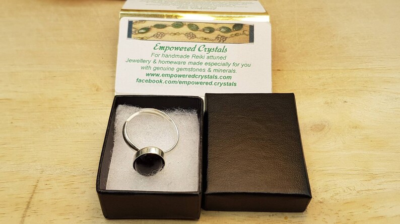 May include: A silver ring with a black gemstone, presented in a black box with a white interior. The box is labelled "Empowered Crystals" with a website and Facebook page.