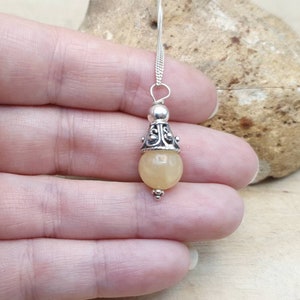 Puede incluir: Un collar con un colgante de plata adornado con una piedra redonda de color amarillo claro y un dise&ntilde;o de plata ornamentado. El colgante cuelga de una cadena de plata.