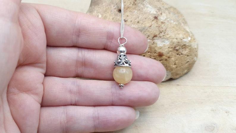 Puede incluir: Un collar con un colgante de plata adornado con una piedra redonda de color amarillo claro y un dise&ntilde;o de plata ornamentado. El colgante cuelga de una cadena de plata.