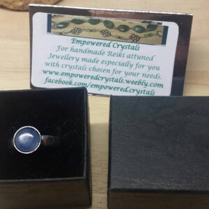 May include: A silver ring with a light blue gemstone set in a bezel setting. The ring is in a black ring box. The box is on a wooden surface. The image also includes a business card with the text "Empowered Crystals" and contact information.
