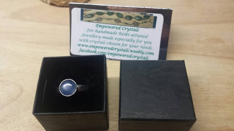 May include: A silver ring with a light blue gemstone set in a bezel setting. The ring is in a black ring box. The box is on a wooden surface. The image also includes a business card with the text "Empowered Crystals" and contact information.