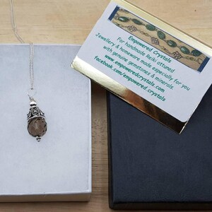 Op de afbeelding: Een zilveren ketting met een bruine edelsteen hanger, gepresenteerd in een open witte doos. Een zwarte doos en een kaart met tekst zijn ook zichtbaar. De kaart leest "Empowered Crystals" en bevat website- en social media-informatie.