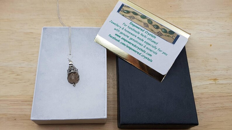 Op de afbeelding: Een zilveren ketting met een bruine edelsteen hanger, gepresenteerd in een open witte doos. Een zwarte doos en een kaart met tekst zijn ook zichtbaar. De kaart leest "Empowered Crystals" en bevat website- en social media-informatie.
