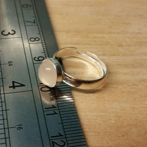 May include: A silver ring with a white oval-shaped stone set in the center. The ring is adjustable and measures approximately 1 inch in diameter.