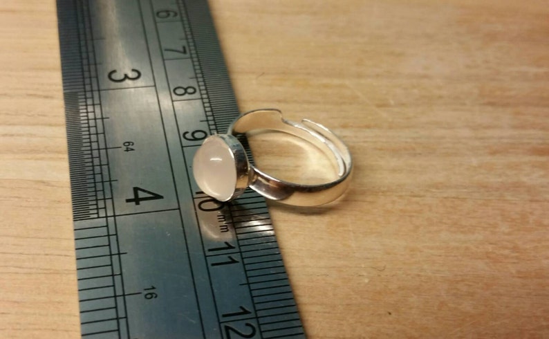 May include: A silver ring with a white oval-shaped stone set in the center. The ring is adjustable and measures approximately 1 inch in diameter.