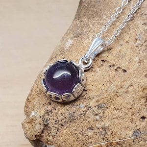 May include: A silver necklace with a purple gemstone pendant. The pendant is set in a silver filigree design.