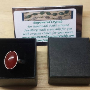 May include: A red carnelian ring in a black ring box. The ring is set in silver and has a smooth, oval-shaped stone. The box is closed and has a white label with the text "Empowered Crystals" and a website address.