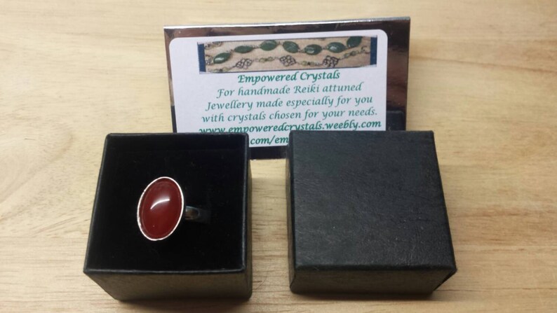 May include: A red carnelian ring in a black ring box. The ring is set in silver and has a smooth, oval-shaped stone. The box is closed and has a white label with the text "Empowered Crystals" and a website address.