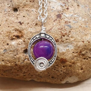 K&ouml;nnte beinhalten: Eine silberne Halskette mit einem Anh&auml;nger aus violettem Stein. Der Anh&auml;nger ist von einem silbernen Rahmen mit einem dekorativen Design umgeben.