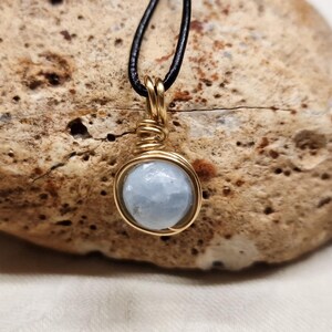May include: A round, pale blue gemstone pendant wrapped in gold-colored wire, suspended from a thin black cord. The stone has a smooth, polished surface and is secured by a coiled wire bail at the top, creating a simple, circular jewelry piece.