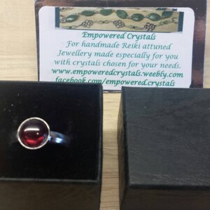 May include: A silver ring with a round, deep red gemstone set in a bezel. The ring is displayed in an open black box, with a business card in the background that reads "Empowered Crystals".