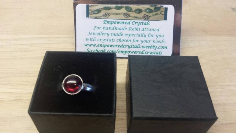 May include: A silver ring with a round, deep red gemstone set in a bezel. The ring is displayed in an open black box, with a business card in the background that reads "Empowered Crystals".