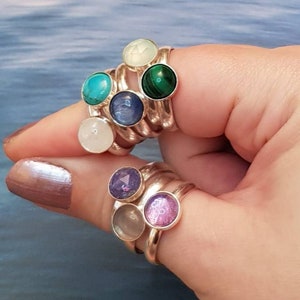 May include: Silver rings with colorful gemstones. The rings feature round gemstones in various colors, including turquoise, green, purple, and pink. The rings are stacked on a finger, showcasing the different gemstone colors and designs.