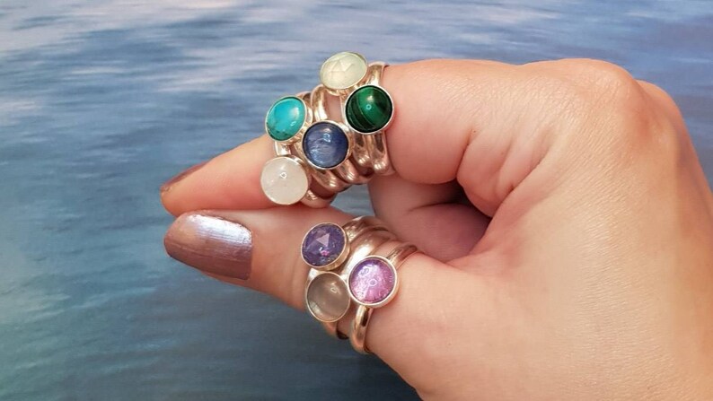 May include: Silver rings with colorful gemstones. The rings feature round gemstones in various colors, including turquoise, green, purple, and pink. The rings are stacked on a finger, showcasing the different gemstone colors and designs.