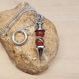 May include: A silver chain with a silver ring and a carnelian crystal pendulum. The pendulum has a silver point and a silver wire wrapped around the carnelian crystal.