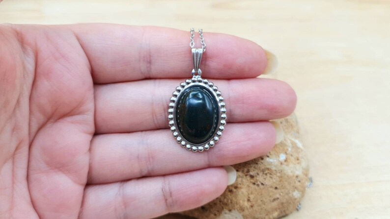 May include: A silver oval pendant necklace with a dark green gemstone. The pendant has a beaded border.