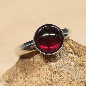 May include: A silver ring with a round, deep red gemstone. The gemstone is set in a bezel setting, and the band is smooth and polished. The ring is displayed on a textured, light brown surface.