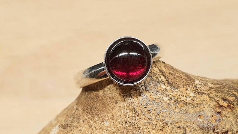 May include: A silver ring with a round, deep red gemstone. The gemstone is set in a bezel setting, and the band is smooth and polished. The ring is displayed on a textured, light brown surface.