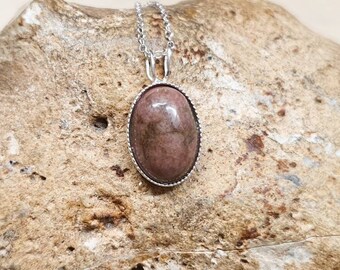 Könnte beinhalten: Eine silberne Kettenkette mit einem ovalen Anhänger in Rosa und Braun. Der Anhänger hat einen silbernen Rand und ist auf einem rauen, braunen Stein platziert.