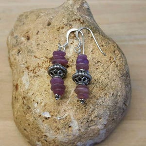 May include: A pair of purple beaded dangle earrings with silver-coloured metal detailing. The earrings are presented on a light brown stone. The earrings feature a stacked bead design with a decorative metal element in the centre.